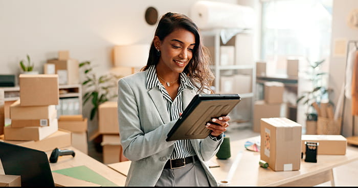 mulher utilizando um tablet em um ambiente de e-commerce pequeno, repleto de caixas de papelão prontas para envio