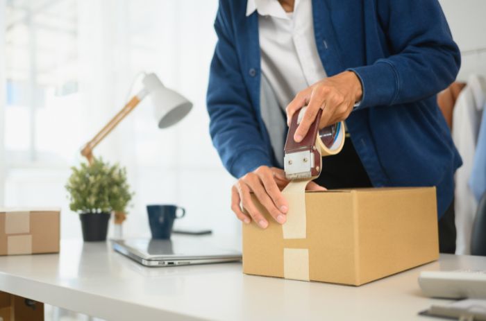 Homem passando fita em uma caixa, dando início a logística de entrega.
